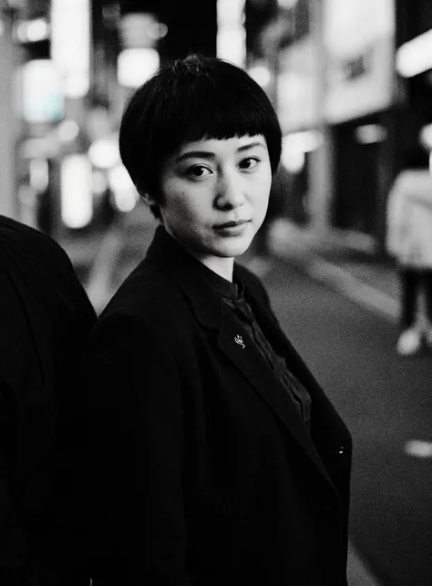 (A black and white photograph:1.1),shot from above,empty Tokyo city street late at night, an Asian woman walking through crowd, looking to viewer, short black hair, wearing a black YSL suit jacket, reflecting the cold, artificial light of the city. The atm...