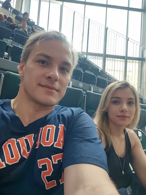 A mature man and woman are seated in a large venue with multiple rows of green seats, hinting at a stadium or arena. 

The man is in the forefront of the photo, capturing a close-up of his face and upper torso. He has fair skin, light blue eyes, and white ...