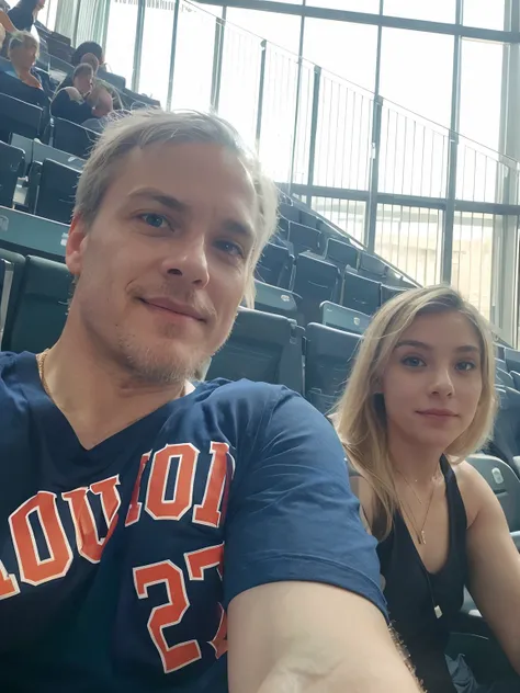 A mature man and woman are seated in a large venue with multiple rows of green seats, hinting at a stadium or arena. 

The man is in the forefront of the photo, capturing a close-up of his face and upper torso. He has fair skin, light blue eyes, and white ...