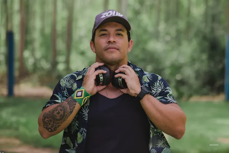 Theres a deejay man whos got a headphone in his hands, pulseira cor verde e amarela no punho direito, Maori tattoo on right forearm, Cap on the head, watch on left wrist, Zouk Writing Cap, blusa florida, camisa preta, In the background there is a forest