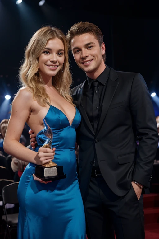 blonde girl singer and guy singer at a music award stand on stage with an award and a smile, Guy in a black suit, Girl in a long blue dress