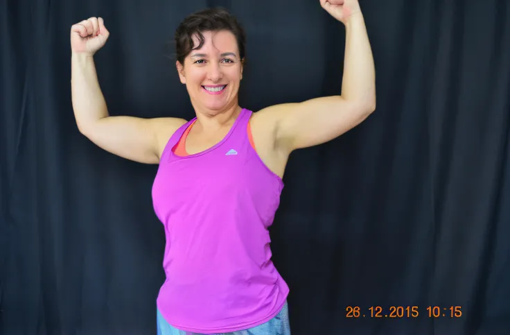 smiling woman in pink blouse flexing arms in front of black backdrop, arms extended, photo from behind, ombro na frente pose, mid 2 0s female, arms to side, 3 0 anos mulher, metade feito, toned arms, Sandra Chevier, mid-transformation, Mulheres de 40 anos,...