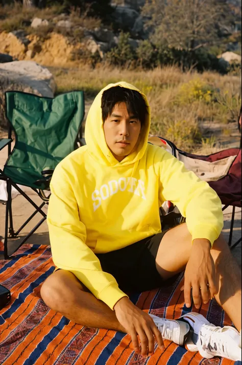 there  a man sitting on a blanket on the ground, wearing a yellow hoodie, Asian man