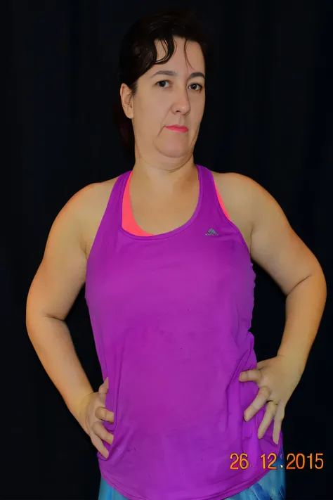 The image shows a woman standing against a black background. She is wearing a purple sports top and has her arms crossed in front of her. Her expression appears neutral and she is looking directly at the camera. The lighting on her face suggests an indoor ...