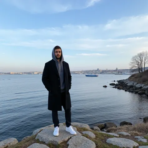 A young man have a light skin tone stands on a rugged cliff overlooking a tranquil bay in Stockholm, Sweden. The scene captures a crisp winter morning with icy blue skies streaked by soft, wispy clouds. The man wears a long black coat with neatly buttoned ...